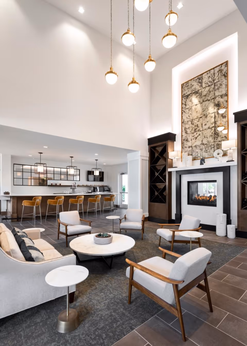 A modern and spacious living room area with a high ceiling, featuring a fireplace with a large decorative mirror above it. The room includes a beige sofa, four light gray armchairs with wooden frames, and round marble-top tables. In the background, there is a kitchen bar area with five mustard-colored bar stools and pendant lights hanging above the counter.