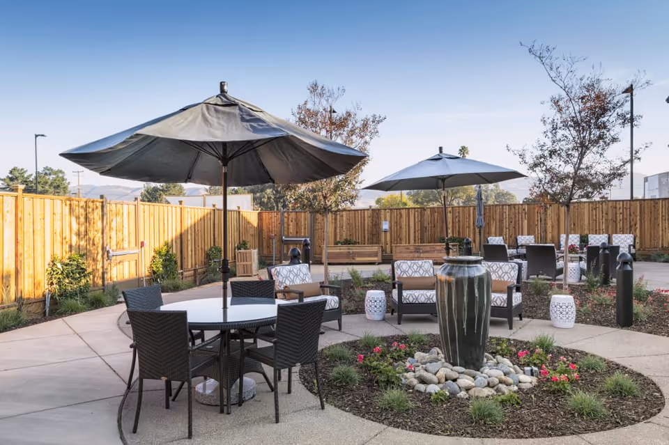 Sunlit outdoor courtyard with round tables and umbrellas, cushioned chairs, a central ceramic fountain surrounded by landscaping and a wooden privacy fence.