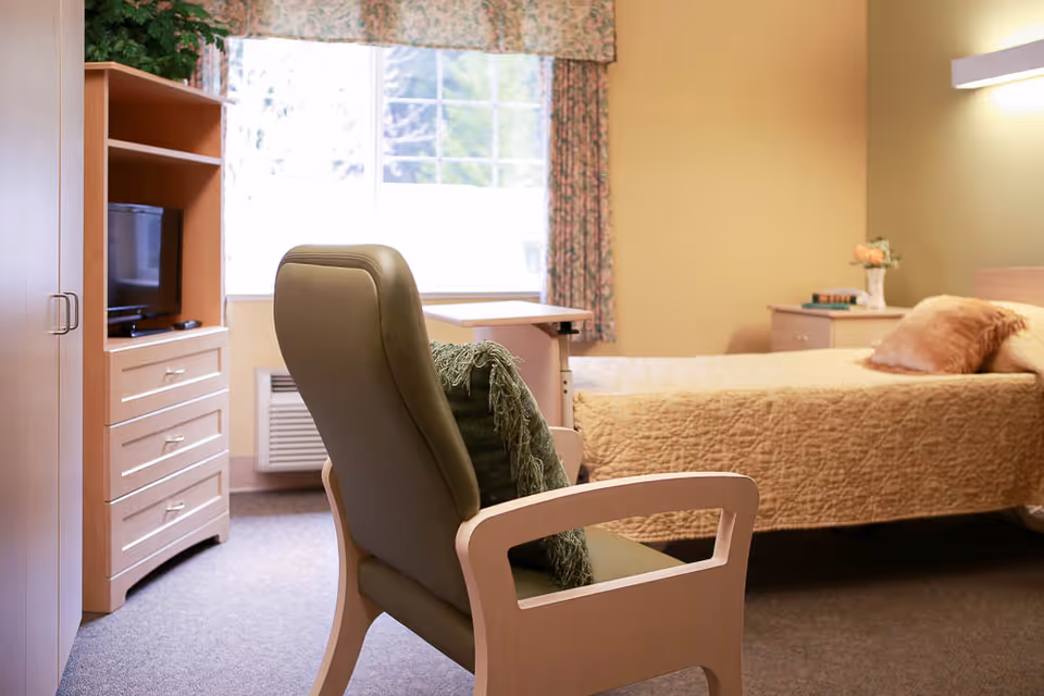 A cozy bedroom in a senior living facility featuring a single bed with a beige quilt and pillows, a wooden nightstand with a small vase of flowers and books, a comfortable armchair with a green cushion, a wooden cabinet with a TV, and a large window with floral curtains letting in natural light.