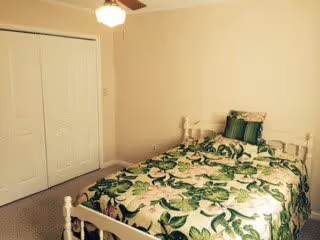 A small bedroom with a white wooden bed frame and a bed covered with a floral green and white bedspread. There are two pillows on the bed, one solid green and one floral. The room has beige walls, a ceiling fan with a light, and a closed white closet with double doors.