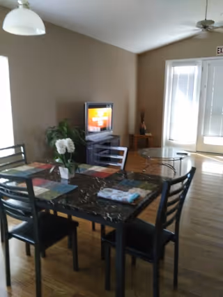 A dining area with a rectangular table covered with a dark marble-like surface and four black chairs. The table has colorful placemats and a vase with white flowers. In the background, there is a TV on a stand, a small side table with a decorative item, and a door with blinds letting in natural light. The floor is wooden, and the walls are painted beige.