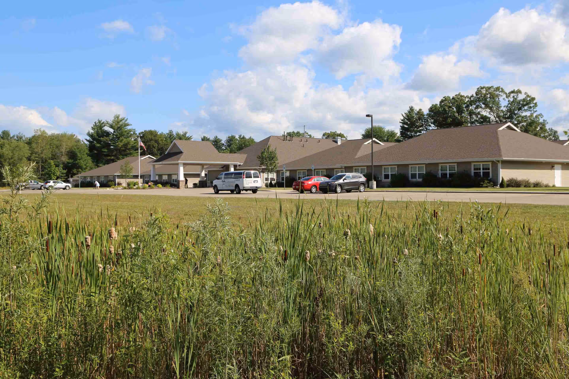 Front exterior of a single-story senior living building with an entrance canopy, several parked cars, and tall grasses in the foreground under a blue sky.