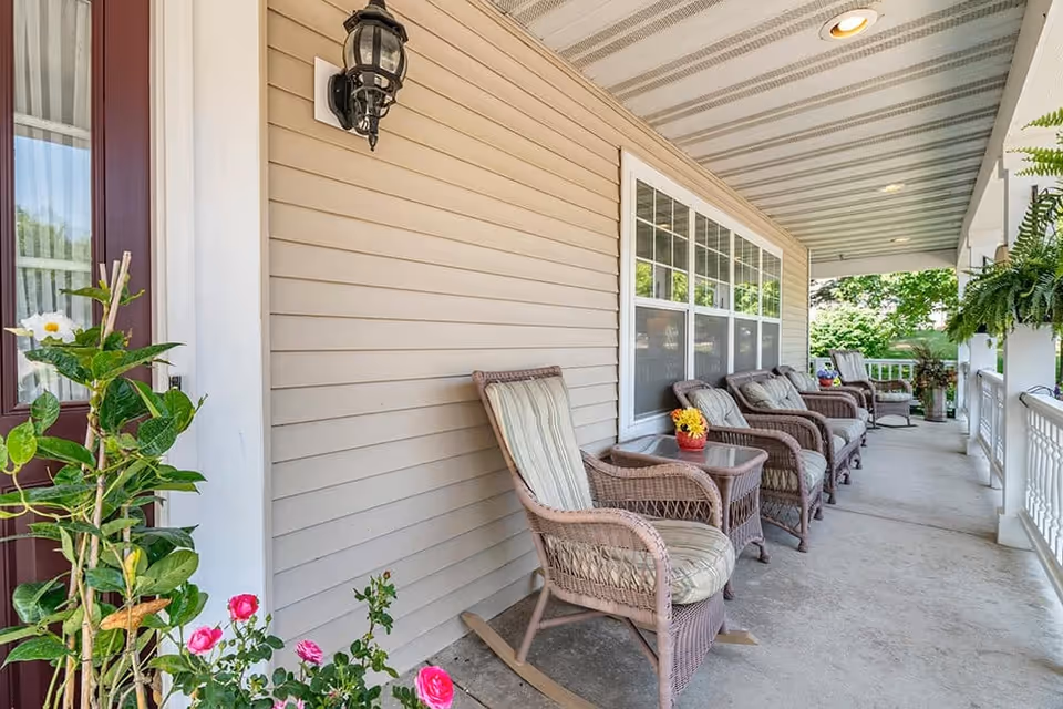 A covered porch area with several cushioned wicker rocking chairs and side tables arranged along the beige siding wall of a building. There are potted plants and flowers near the entrance door and hanging ferns along the porch railing. The porch overlooks a green outdoor area with trees.