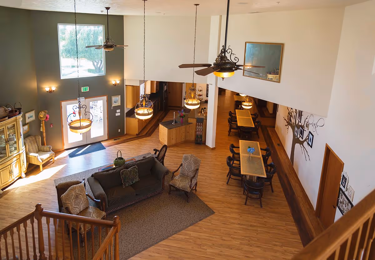 Interior view of a senior living facility common area with wooden flooring, a seating area with a sofa and armchairs on a rug, dining tables with chairs, ceiling fans with lights, and large windows letting in natural light.