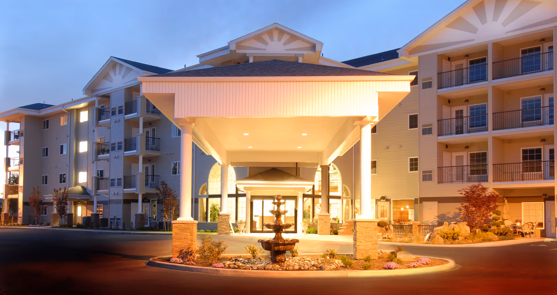 Exterior view of a senior living community building at dusk with a well-lit entrance featuring a covered driveway supported by columns and a decorative fountain in the center of a landscaped roundabout.