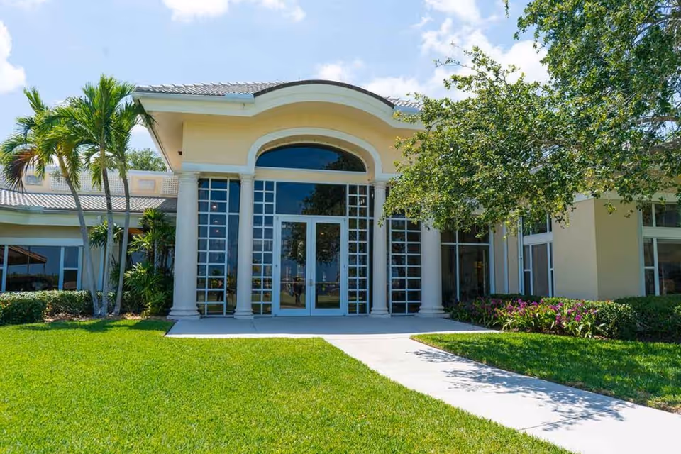 Front exterior view of a building with large glass doors and windows framed by white columns, surrounded by green grass, palm trees, and other plants under a blue sky with some clouds.