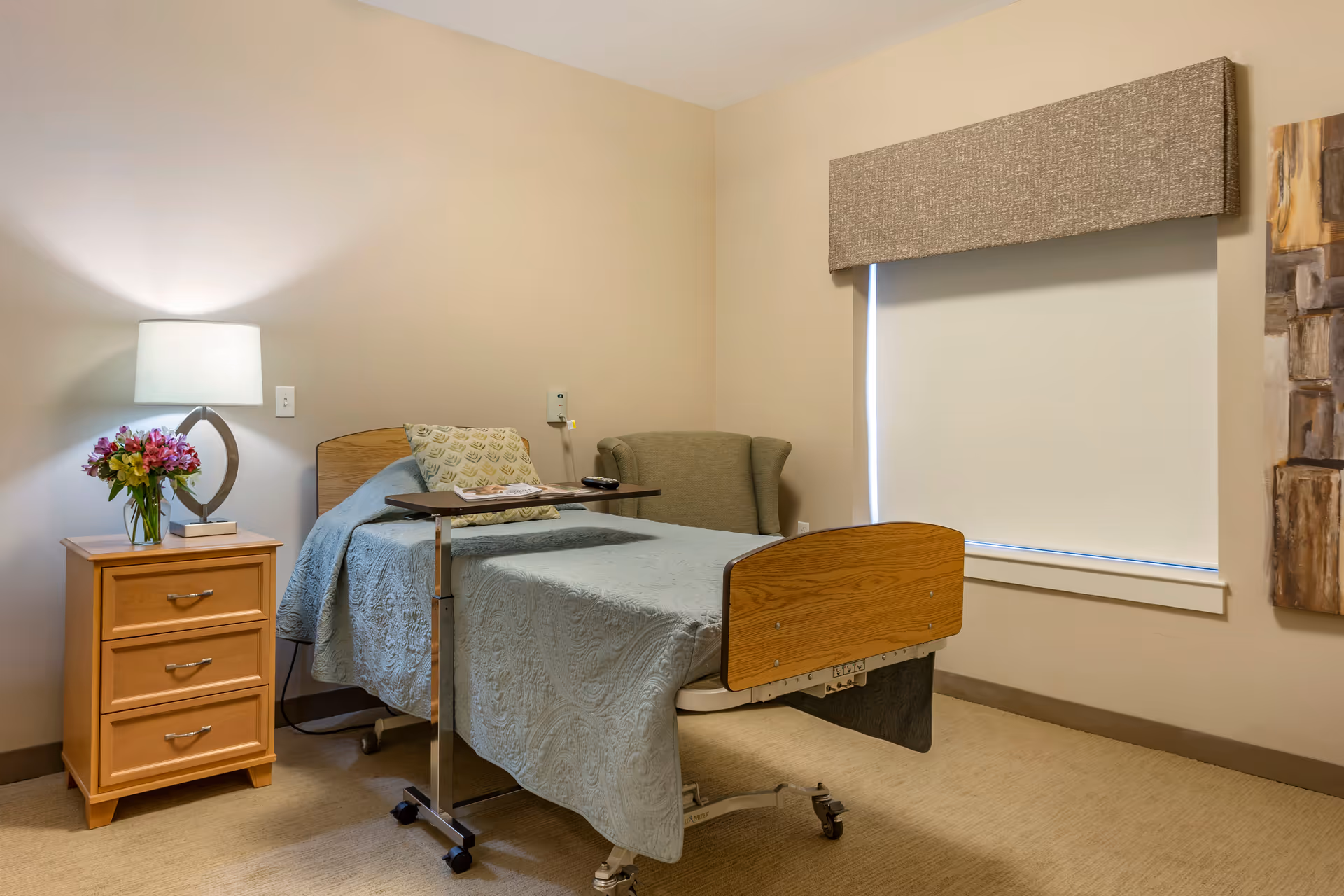 A senior living facility bedroom with a hospital-style bed covered in a light blue quilt, a small wooden nightstand with a lamp and a vase of flowers, a cushioned armchair, and a window with a closed blind and fabric valance.