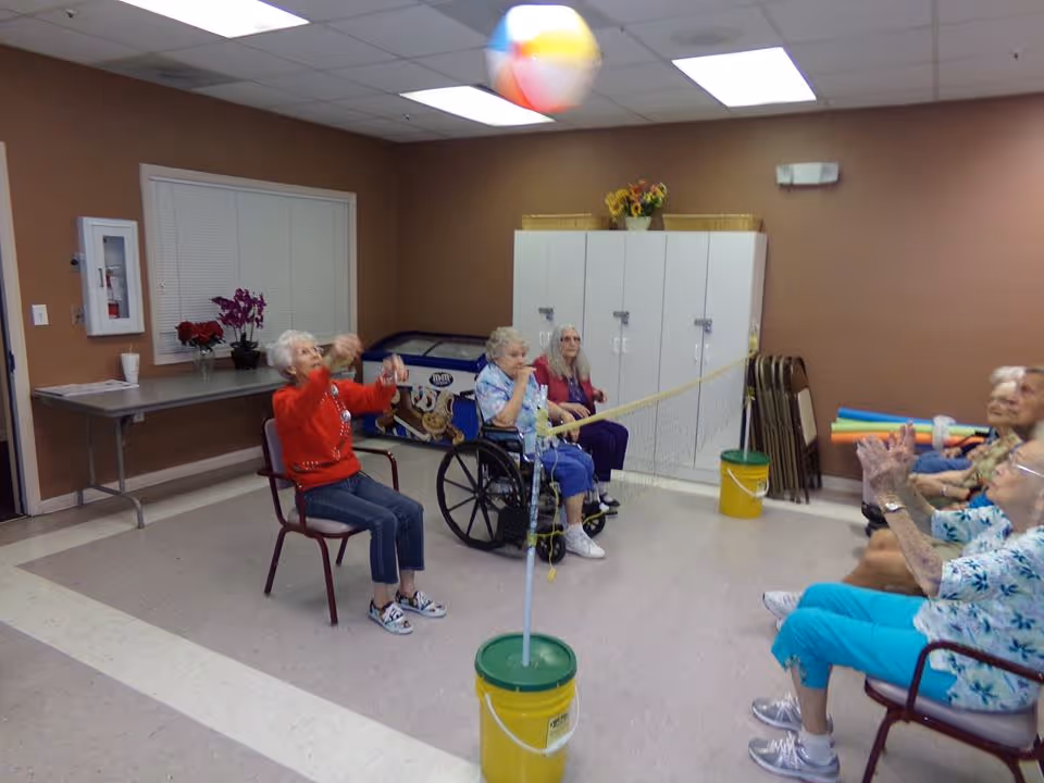 A group of elderly women seated in chairs and a wheelchair playing a seated volleyball game indoors with a beach ball and a makeshift net made from a rope tied between two yellow buckets.