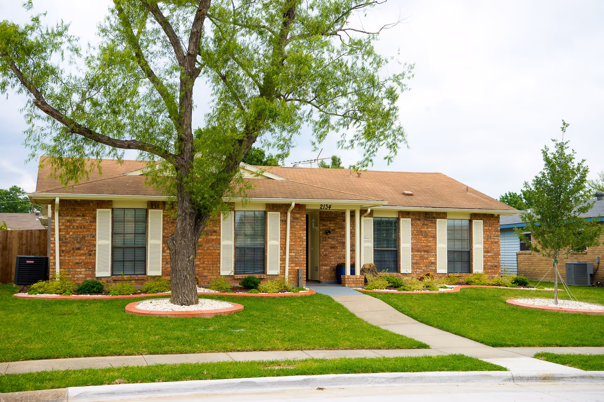 Single-story brick house with a brown shingle roof, white window shutters, and a front porch. The house is surrounded by a well-maintained green lawn with a large tree in the front yard and smaller plants along the foundation. A concrete sidewalk leads to the front door.