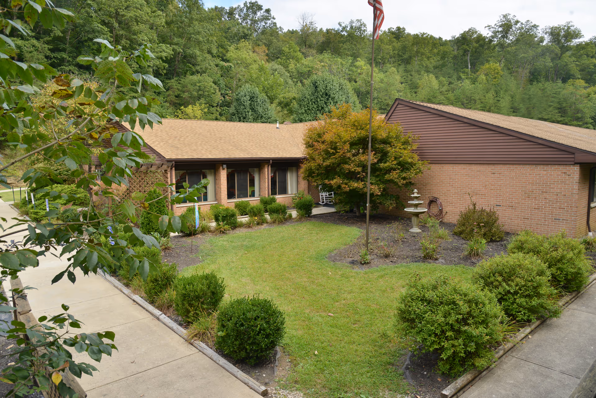 Single-story brick senior living building with a grassy courtyard, flagpole, shrubs and trees.