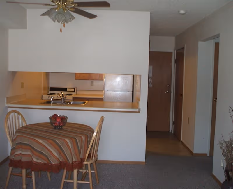 Interior view of a small dining area and kitchen in a senior living facility. The dining area has a round wooden table covered with a striped tablecloth and two wooden chairs. On the table is a bowl of red apples. The kitchen is visible through a pass-through window and includes a sink, stove, and refrigerator. A ceiling fan with a light fixture is mounted above the dining area. There is a hallway leading to other rooms with wooden doors.