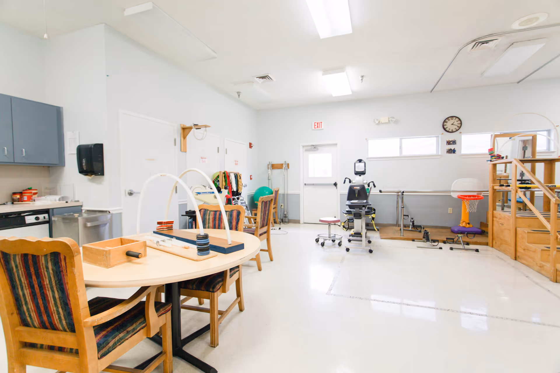 A bright therapy or activity room in a senior living facility with a table and chairs, therapy equipment, exercise bike, parallel bars, and a small basketball hoop. The room has white walls, a clock, and a door with an exit sign above it.