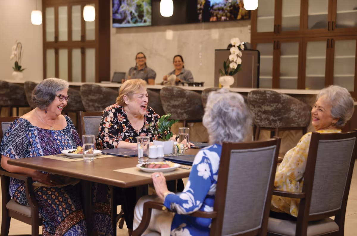 Four elderly women laughing around a dining table in a well-appointed communal dining area with staff at a counter in the background.