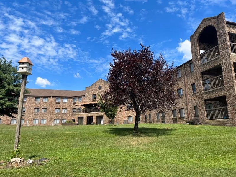 A large brick senior living facility building with multiple windows and balconies surrounding a green lawn. In the foreground, there is a tree with dark red leaves and a tall wooden birdhouse on a pole. The sky is blue with scattered clouds.
