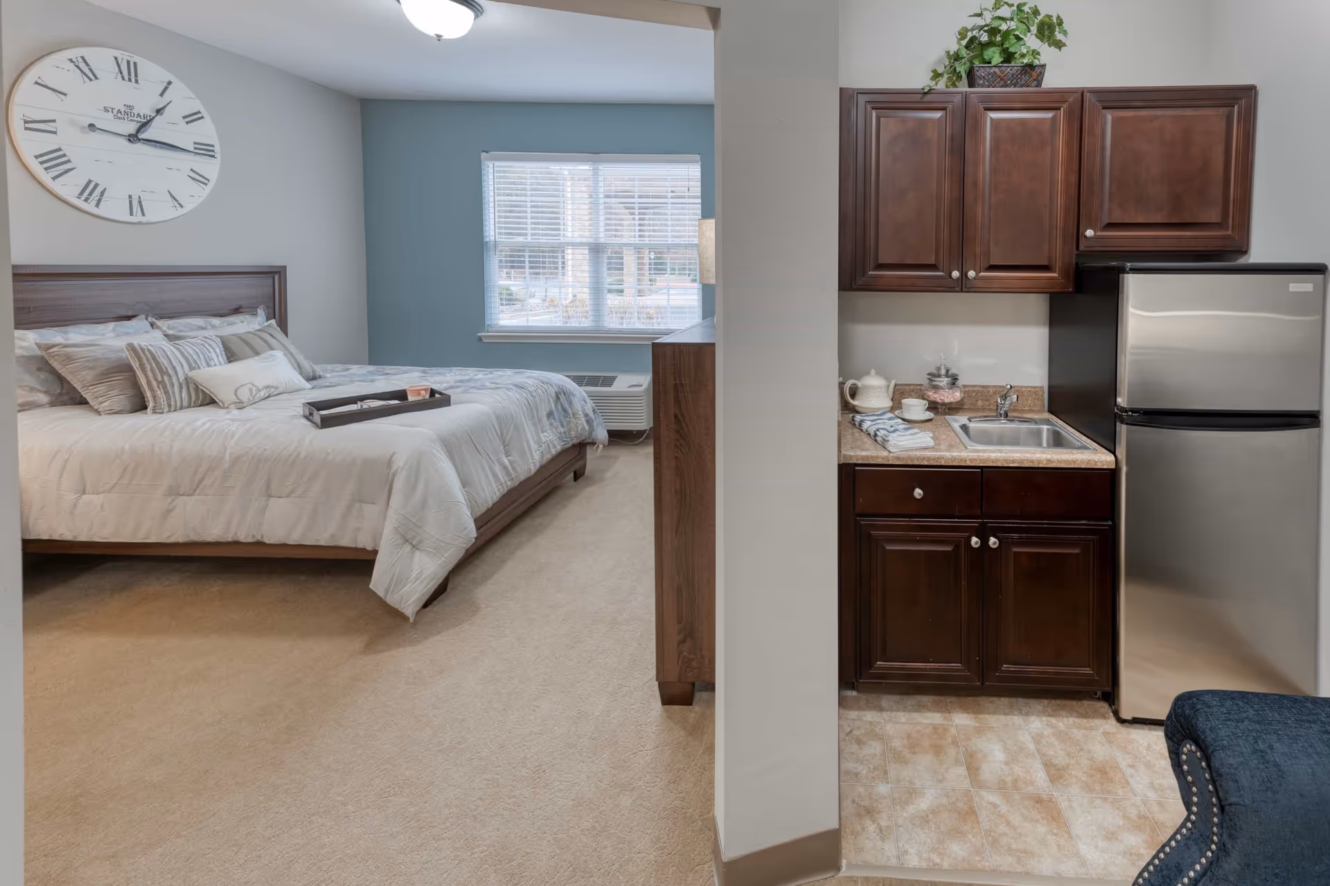 View of a senior living facility room showing a bedroom with a large bed, decorative pillows, a wall clock, and a window with blinds. Adjacent to the bedroom is a small kitchenette with dark wooden cabinets, a sink, a stainless steel refrigerator, and a countertop with a teapot and cups.