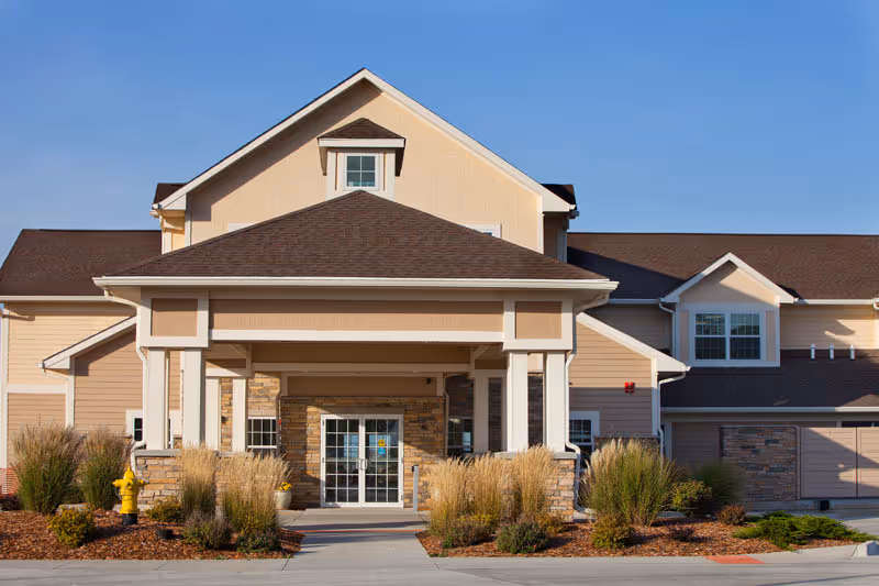 Front entrance of a beige senior living building with a covered porte-cochere, glass double doors, and landscaped grasses.