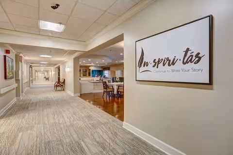 A long, well-lit hallway in a retirement community with carpeted floors and beige walls. On the right wall, there is a large framed sign that reads 'Inspiritas Continue to Write Your Story.' The hallway has several chairs along the walls and leads to an open area with a desk and chairs, featuring wooden flooring.