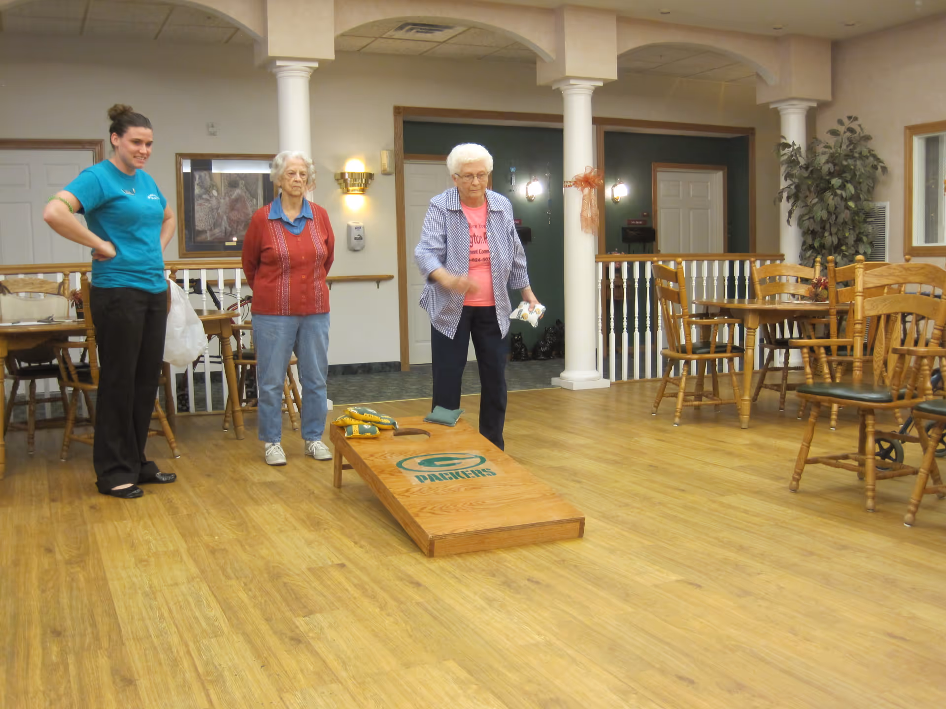 Three women in a senior living facility common area playing a game of cornhole. One elderly woman is tossing a bean bag towards a wooden cornhole board with a Green Bay Packers logo, while another elderly woman and a younger woman watch. The room has wooden floors, wooden tables and chairs, white columns, and framed artwork on the walls.
