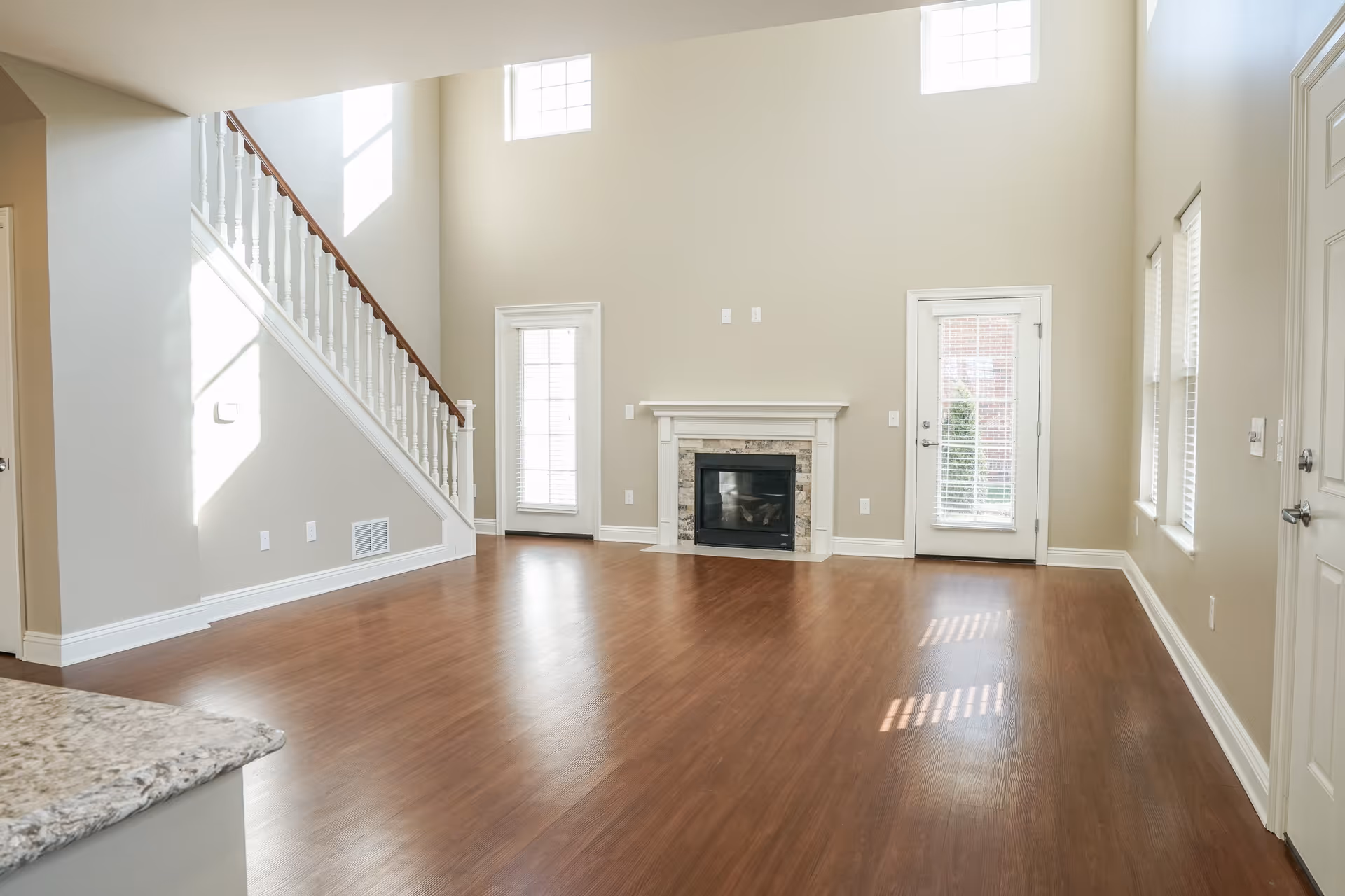 Empty living room with wooden flooring, a white fireplace with stone accents, two doors with glass panes and blinds, three windows with blinds, and a staircase with white railing leading to the upper floor. The walls are painted beige and the room is well-lit with natural light.
