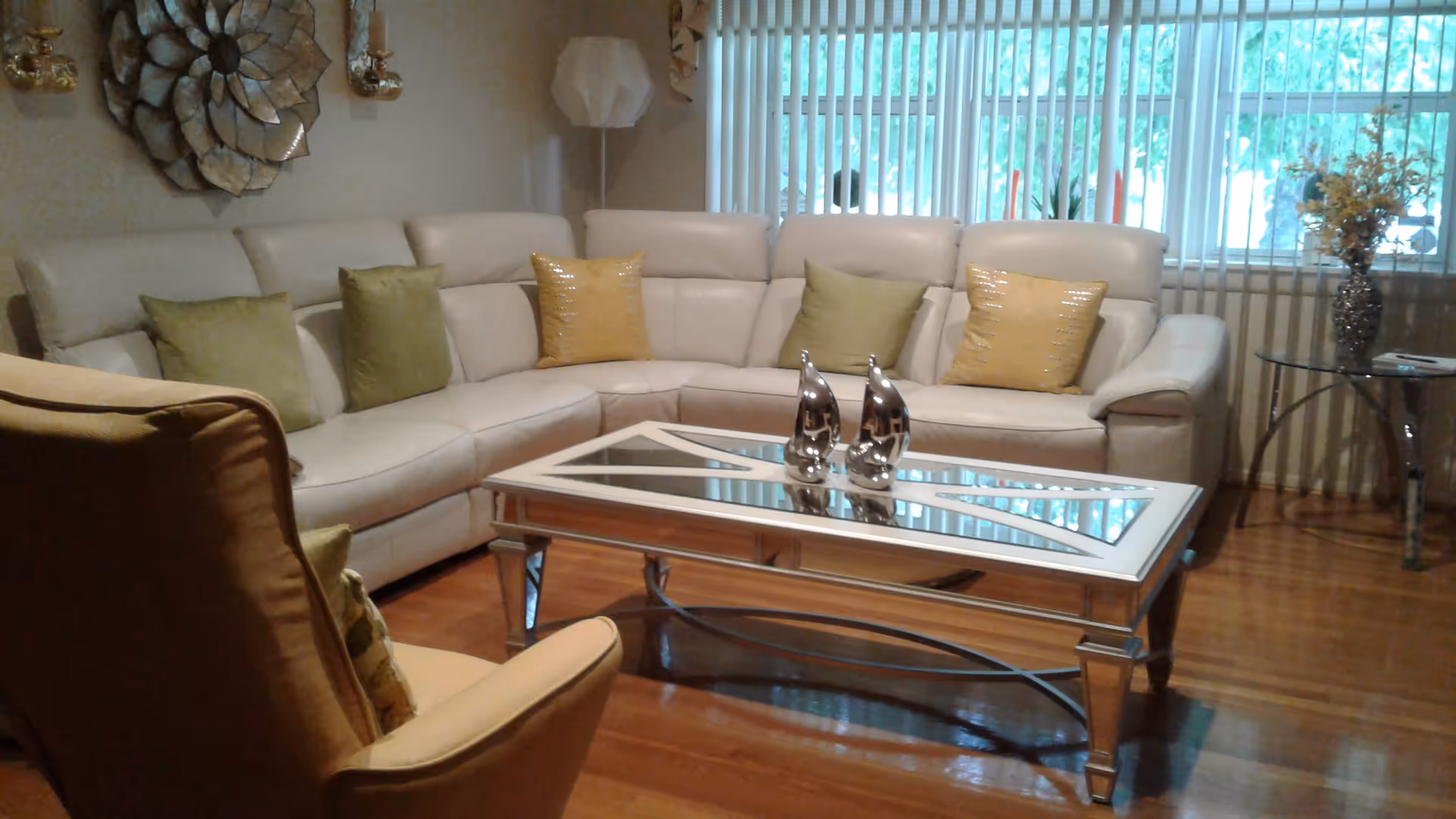 A cozy living room with a large beige sectional sofa adorned with green and yellow pillows. In front of the sofa is a glass-top coffee table with two silver decorative sculptures. There is a beige armchair in the foreground, a decorative wall piece, and a floor lamp in the corner. A large window with vertical blinds lets in natural light, and a small round glass side table with a vase of flowers is next to the sofa.