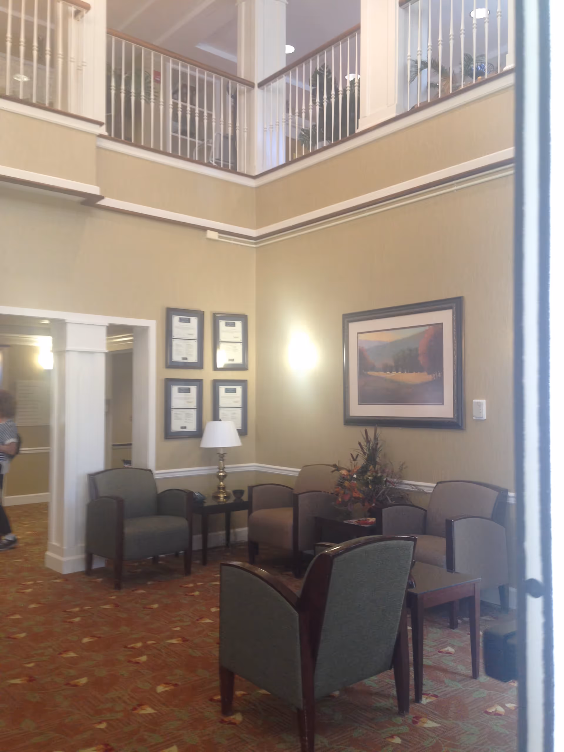 A seating area inside a senior living facility with several upholstered chairs arranged around small tables. The walls are beige with white trim, decorated with framed certificates and a landscape painting. There is a lamp on one of the tables and a floral arrangement on another. The area has a high ceiling with a balcony railing visible above.