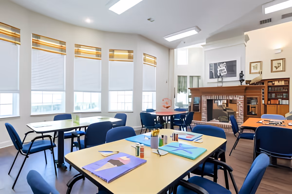 A bright room with several tables and blue chairs arranged around them. The tables have colorful papers, art supplies, and paint bottles on them. Large windows with white blinds let in natural light. In the background, there is a fireplace with a mirror above it and bookshelves on the side, creating a cozy atmosphere.
