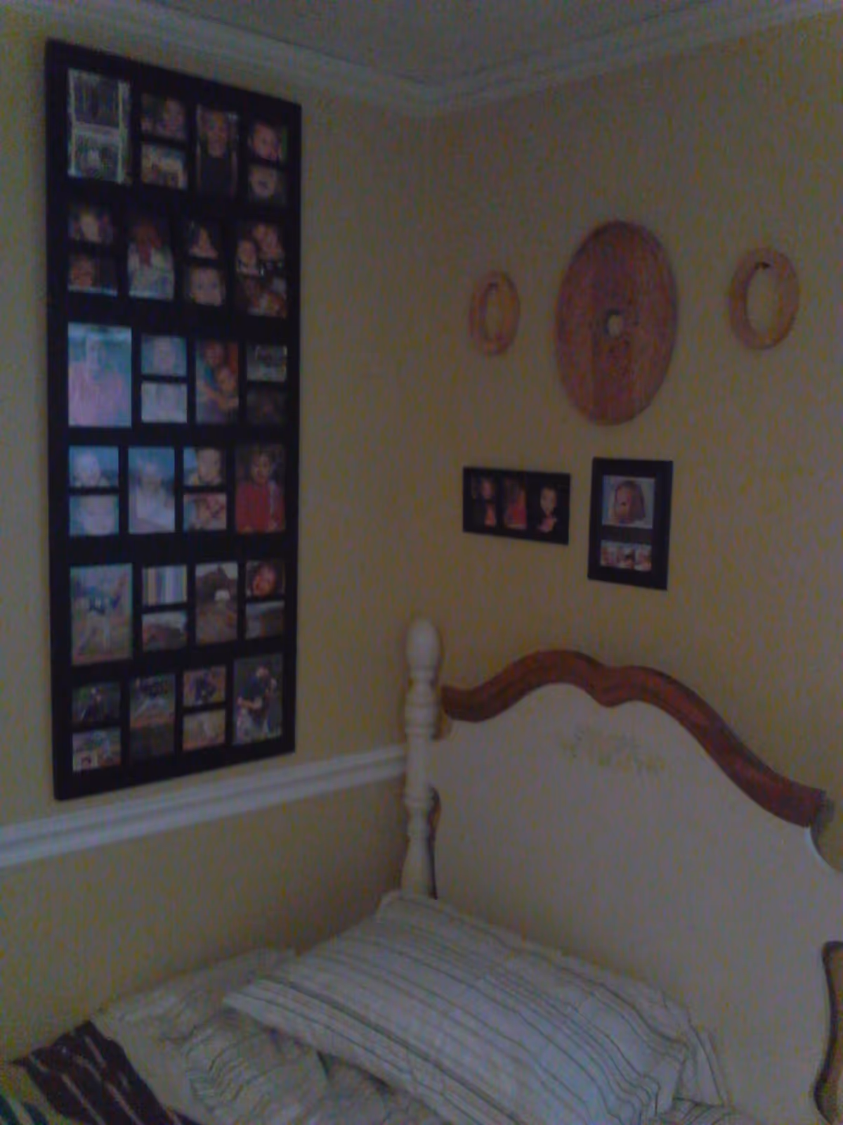 A bedroom corner with a white wooden bed frame and striped bedding. The walls are painted light yellow and decorated with multiple photo frames containing various pictures and three circular wall hangings above the bed.