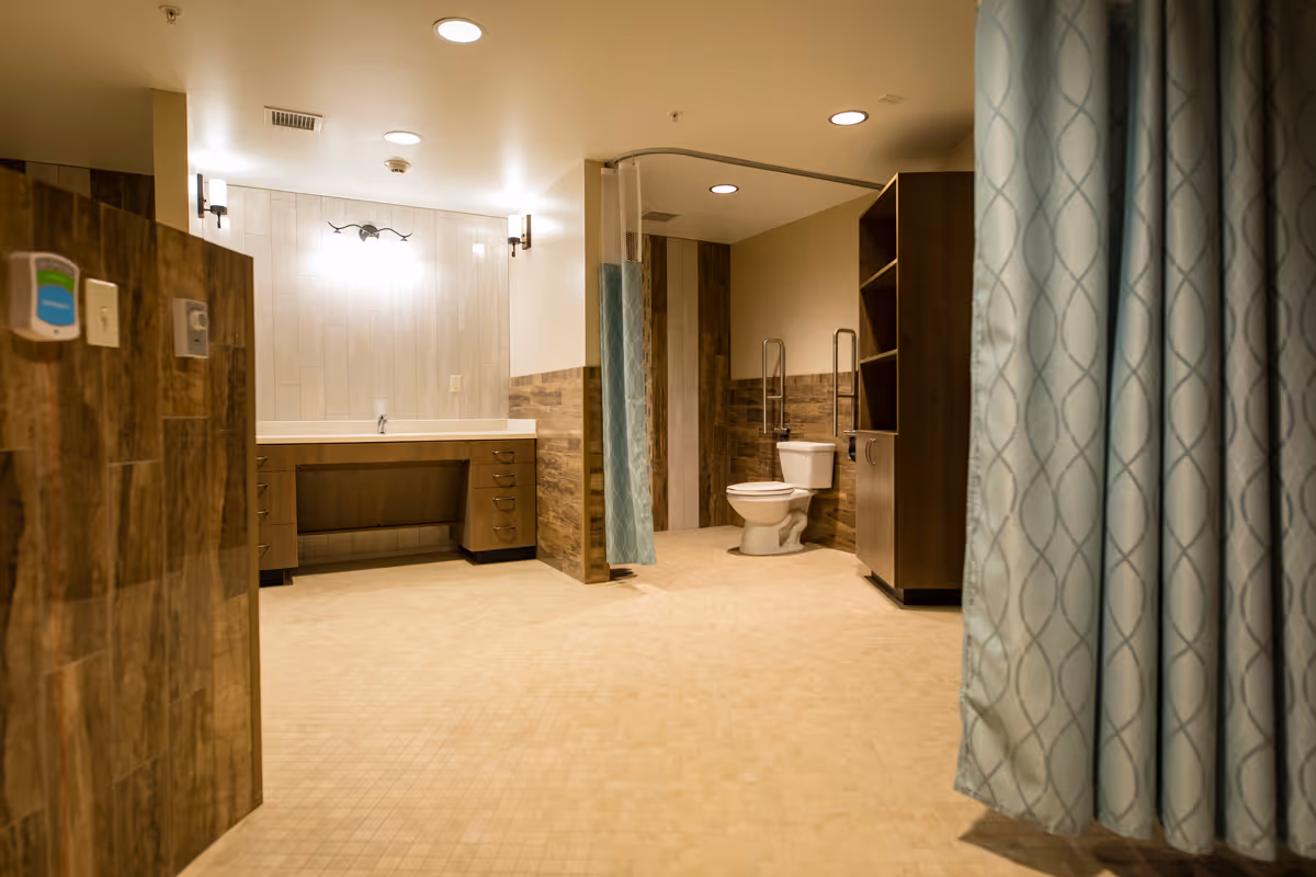 Spacious senior living bathroom with beige tiled floor and walls partially covered with wood paneling. There is a white toilet with grab bars, a large vanity with a sink and multiple drawers, and a blue patterned privacy curtain. The room is well-lit with ceiling lights and wall sconces.