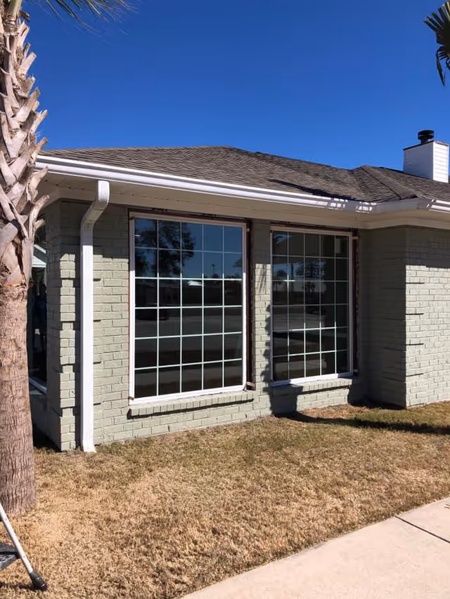 Exterior view of a single-story building with light green brick walls, two large grid-style windows, a sloped shingle roof, and a palm tree on the left side. The ground is covered with dry grass and a concrete walkway is visible on the right.