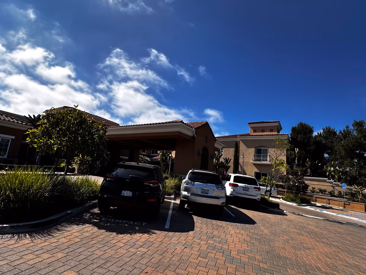 Front entrance and parking area of a senior living facility with several cars parked under a bright blue sky.