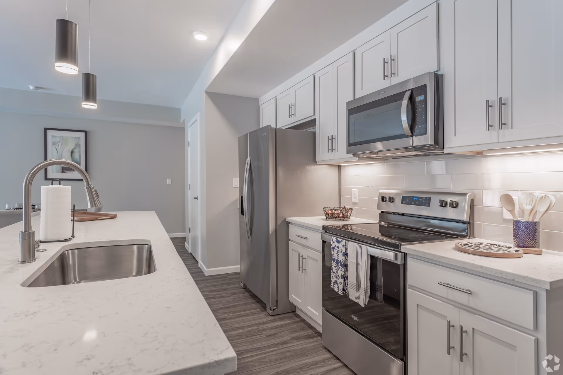 Modern kitchen with white cabinets, stainless steel refrigerator, stove, microwave, and a large marble countertop island with a sink and paper towel holder. Two pendant lights hang above the island, and there is a framed artwork on the wall in the background.