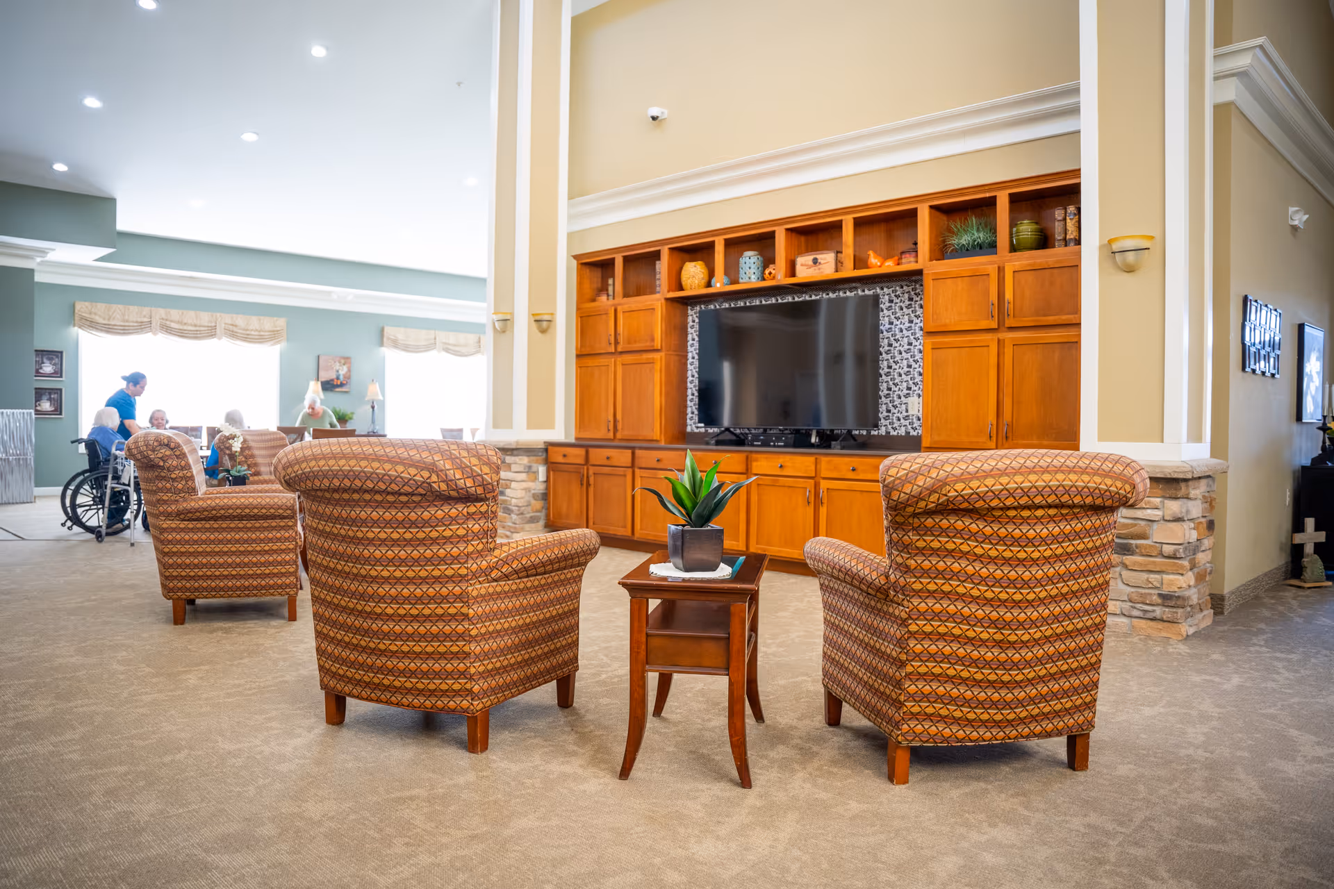A cozy living room area in a senior living facility with three patterned armchairs arranged around a small wooden table with a potted plant. In the background, there is a large wooden entertainment center with a flat-screen TV and decorative items on the shelves. To the left, several seniors and a caregiver are visible near large windows letting in natural light.