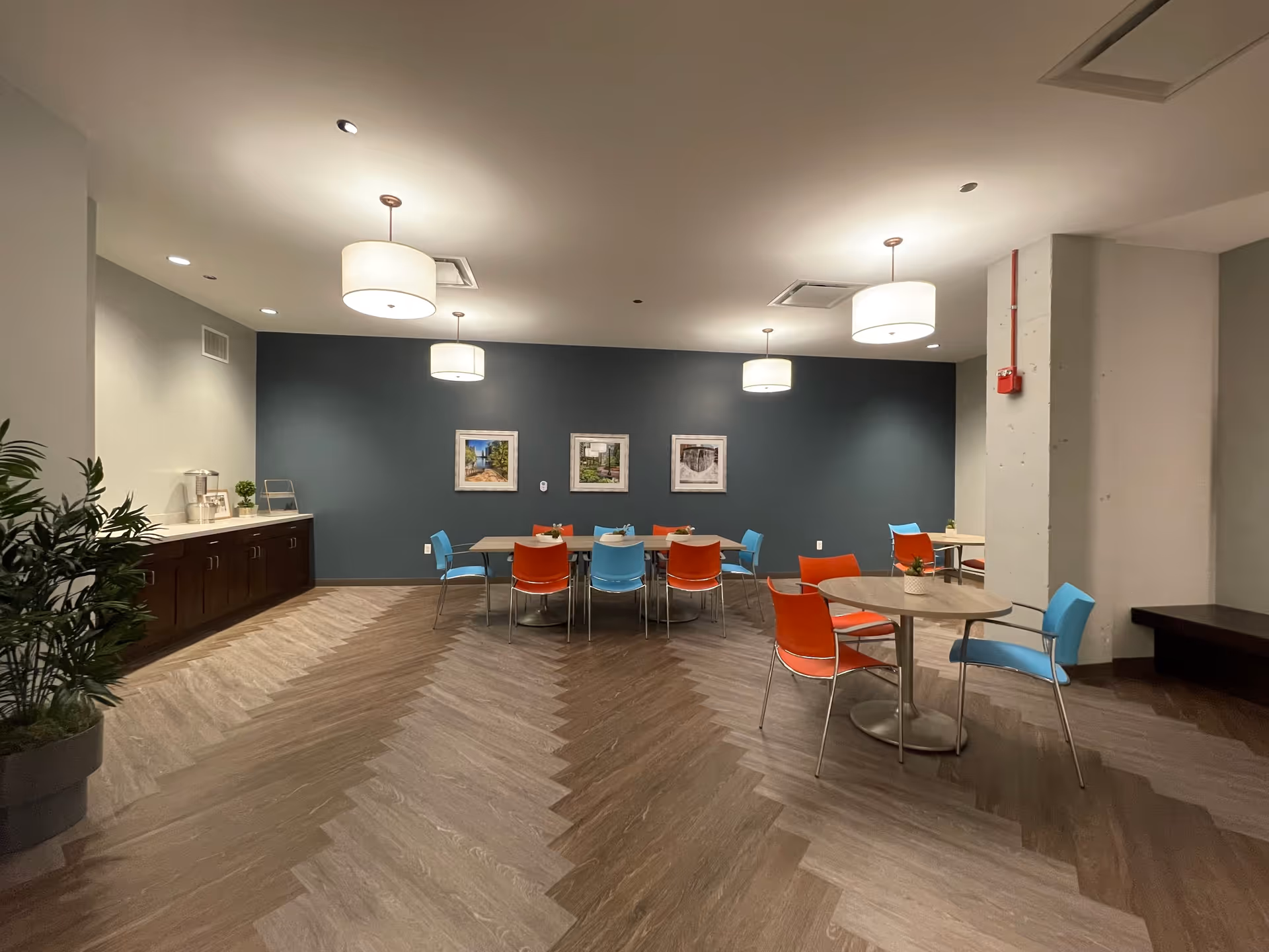 A modern dining area with a dark accent wall featuring three framed pictures. The room has multiple round and rectangular tables surrounded by red and blue chairs. There are three large ceiling lights and a side counter with a water dispenser and small plants. The floor has a chevron pattern in light and dark wood tones.