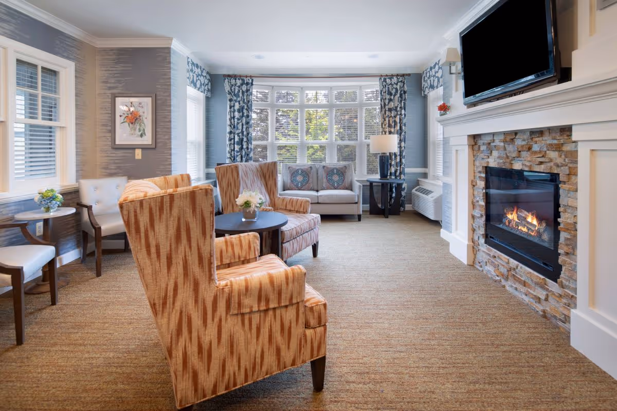 Sunlit communal living room with upholstered chairs and sofa, a stone fireplace with mounted TV, and a large bay window.