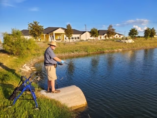 An elderly man wearing a hat and gloves is fishing by the edge of a pond near a grassy area. A blue walker is placed nearby on the grass. In the background, there is a large single-story building with a covered entrance and several trees under a clear blue sky.
