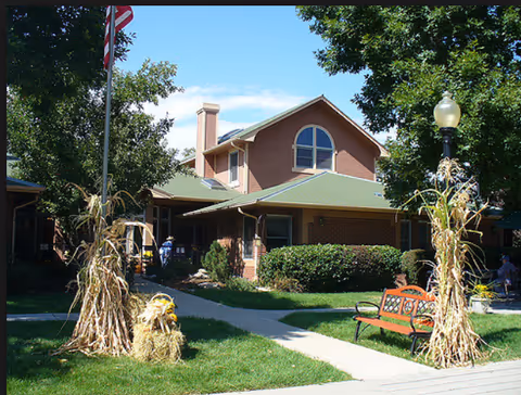 Exterior view of a two-story brick building with a green roof, surrounded by trees and bushes. There is a paved walkway leading to the entrance, an American flag on a pole, and decorative hay bales with dried corn stalks on either side of the walkway. A red bench is placed near a lamppost on the right side.