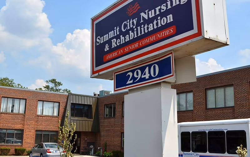Exterior view of Summit City Nursing & Rehabilitation building with a large sign displaying the facility name and address number 2940. A white shuttle bus and a silver car are parked in front of the brick building under a partly cloudy sky.