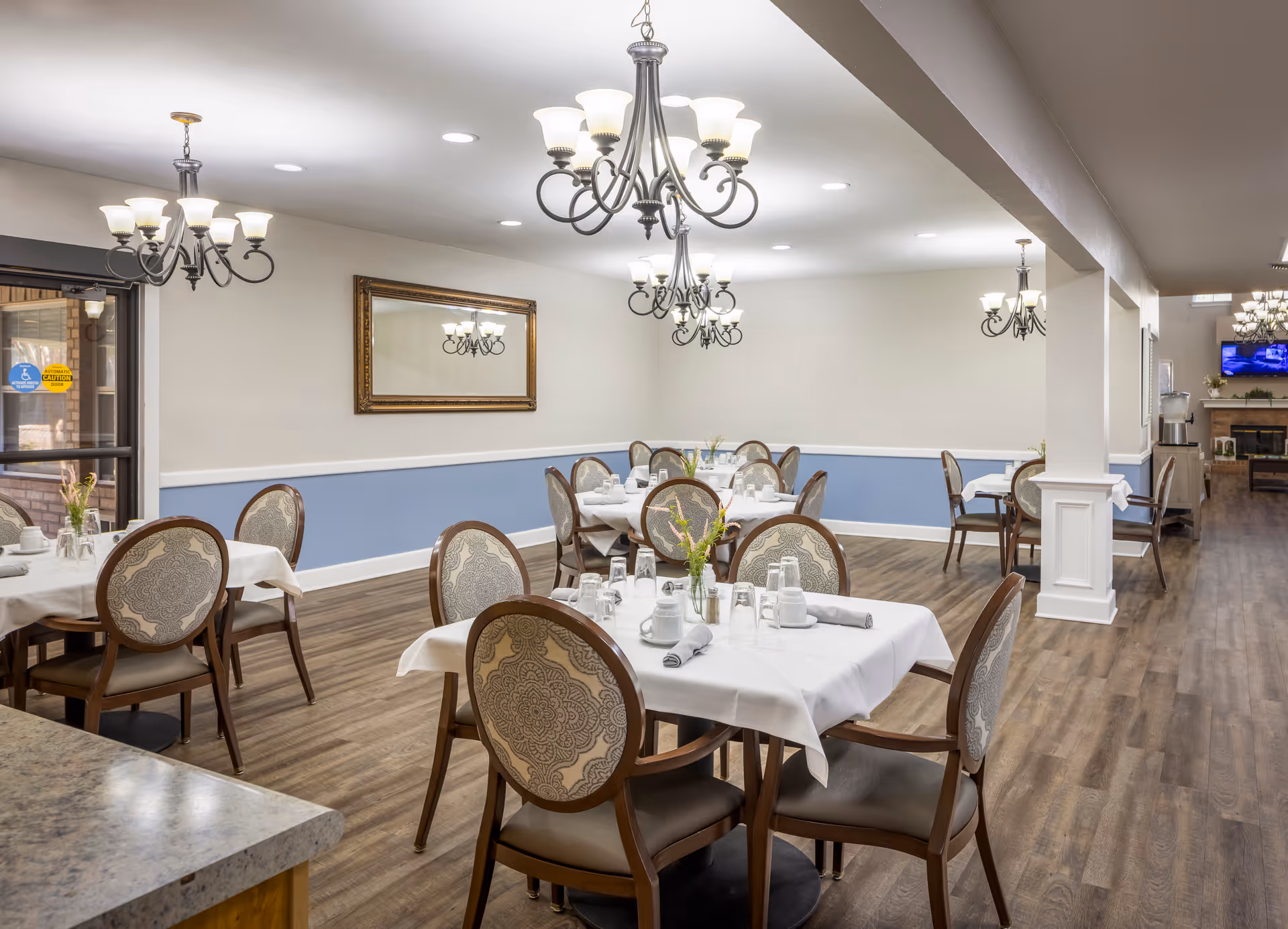 A dining room with several tables covered in white tablecloths, each set with cups, glasses, and napkins. The room has wooden floors, light blue and white walls, ornate chandeliers hanging from the ceiling, and a large mirror on one wall. There is a doorway leading to another room with a fireplace and a TV.