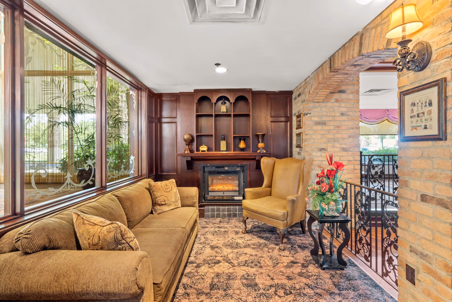 Cozy sitting area with a brown sofa and a tan armchair facing a fireplace with wooden shelves above it. The room features a large window with decorative glass on the left and a brick archway on the right leading to another area. A small table with a floral arrangement is placed next to the armchair, and the floor is covered with a patterned carpet.