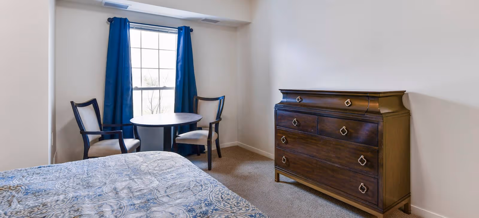 Bright bedroom with a bed, a wooden dresser, and a small round table with two chairs by a window with blue curtains.