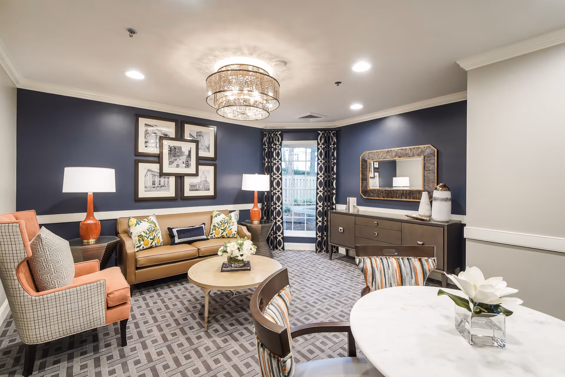 A cozy and stylish living room with a tan leather sofa adorned with decorative pillows, two orange and beige armchairs, a round coffee table with a flower arrangement, and a round dining table with chairs. The walls are painted navy blue with white trim, decorated with framed black and white photos and a large mirror. Two table lamps with orange bases provide warm lighting, and a modern chandelier hangs from the ceiling. A glass door with patterned curtains leads outside.