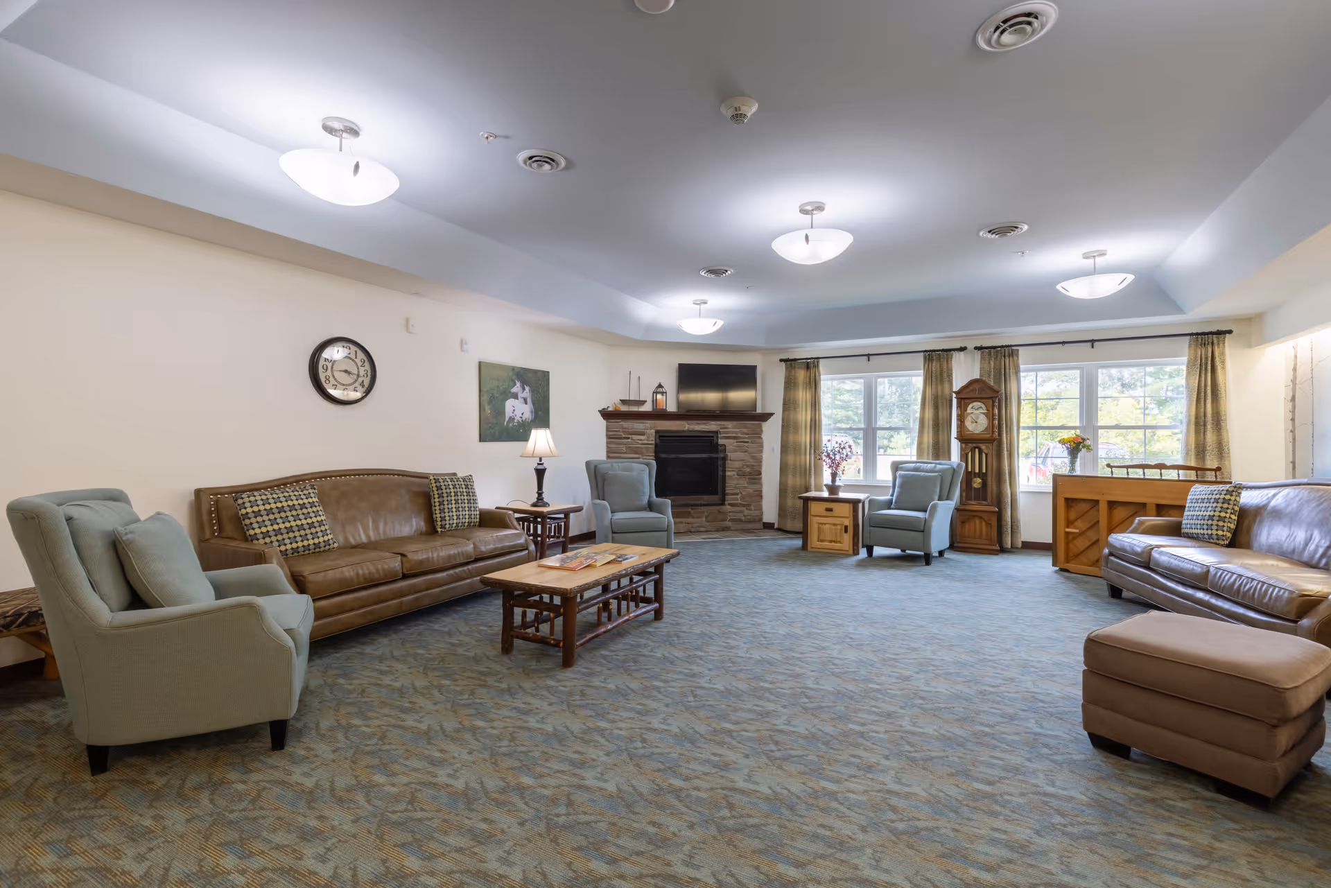A spacious senior living facility common room with beige walls and blue patterned carpet. The room features two brown leather sofas, two light blue armchairs, a wooden coffee table, a side table with a lamp, a stone fireplace with a TV mounted above it, large windows with curtains, a grandfather clock, and a piano with a flower vase on top.