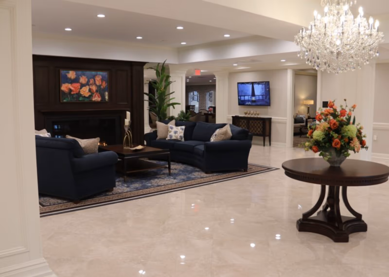 A spacious and elegant living room area with polished marble floors, a dark wood round table with a large floral arrangement, a crystal chandelier hanging from the ceiling, and a seating area with navy blue sofas and a dark wood coffee table in front of a fireplace with a floral painting above it. A flat-screen TV is mounted on the wall in the background.