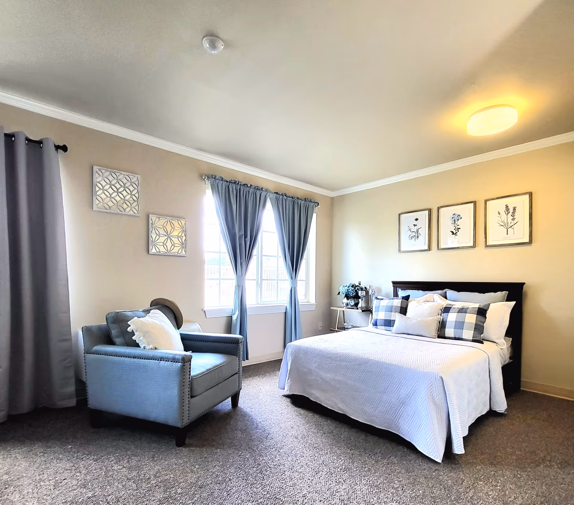 A bright and neatly arranged bedroom with a large bed covered in white bedding and multiple pillows, including checkered ones. There is a comfortable armchair with a pillow and a hat on it near a window with blue curtains. The walls are decorated with three framed botanical prints above the bed and two decorative wall pieces near the window. The room has beige walls, carpeted floor, and a ceiling light.