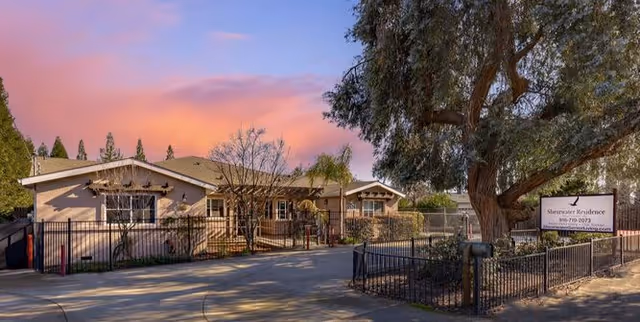 Exterior view of Shearwater Senior Living facility at sunset with a large tree and a sign displaying the facility name and contact number near the entrance gate.
