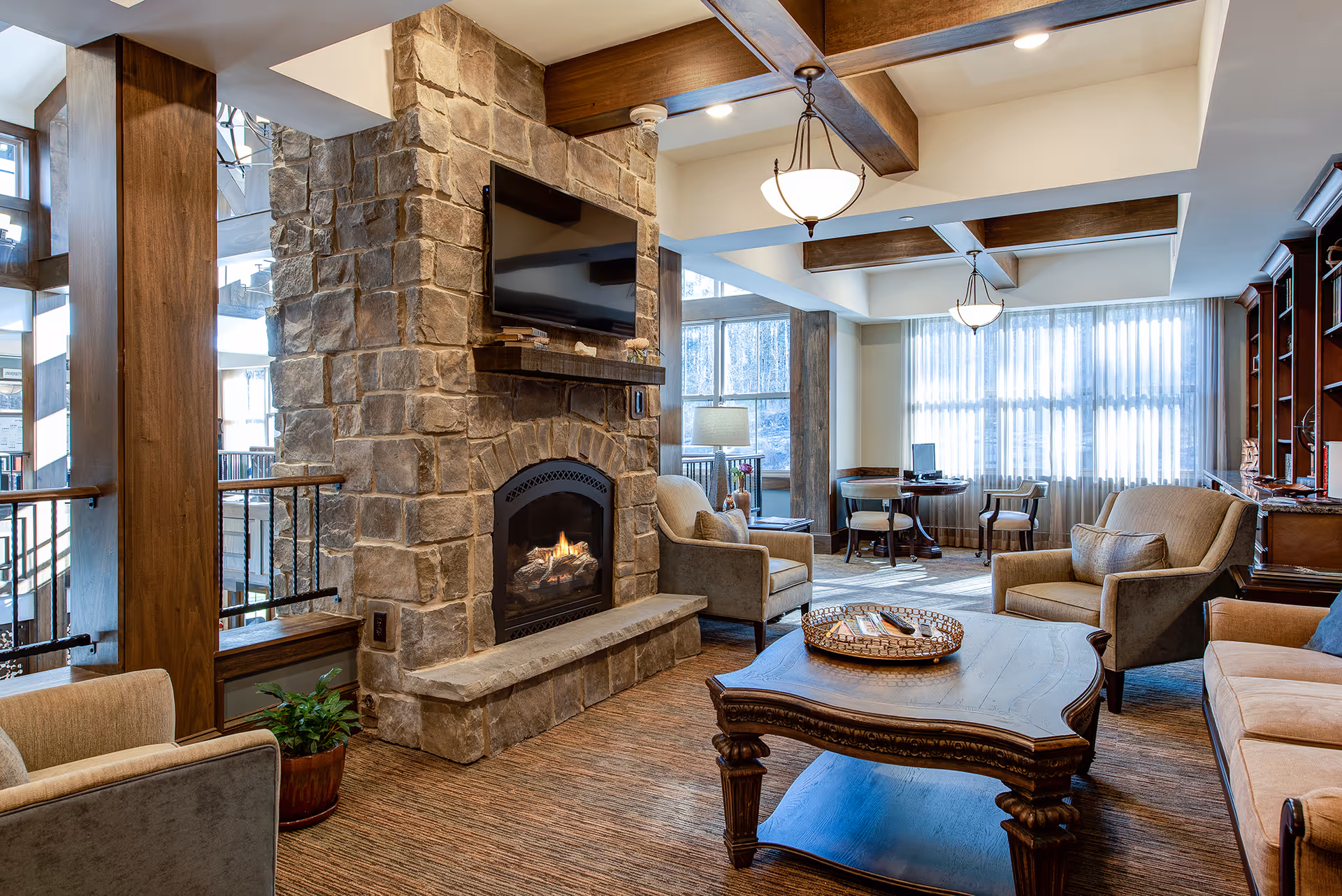 A cozy senior living lounge with a stone fireplace and mounted TV, upholstered chairs and sofa around a wooden coffee table, and large windows letting in natural light.