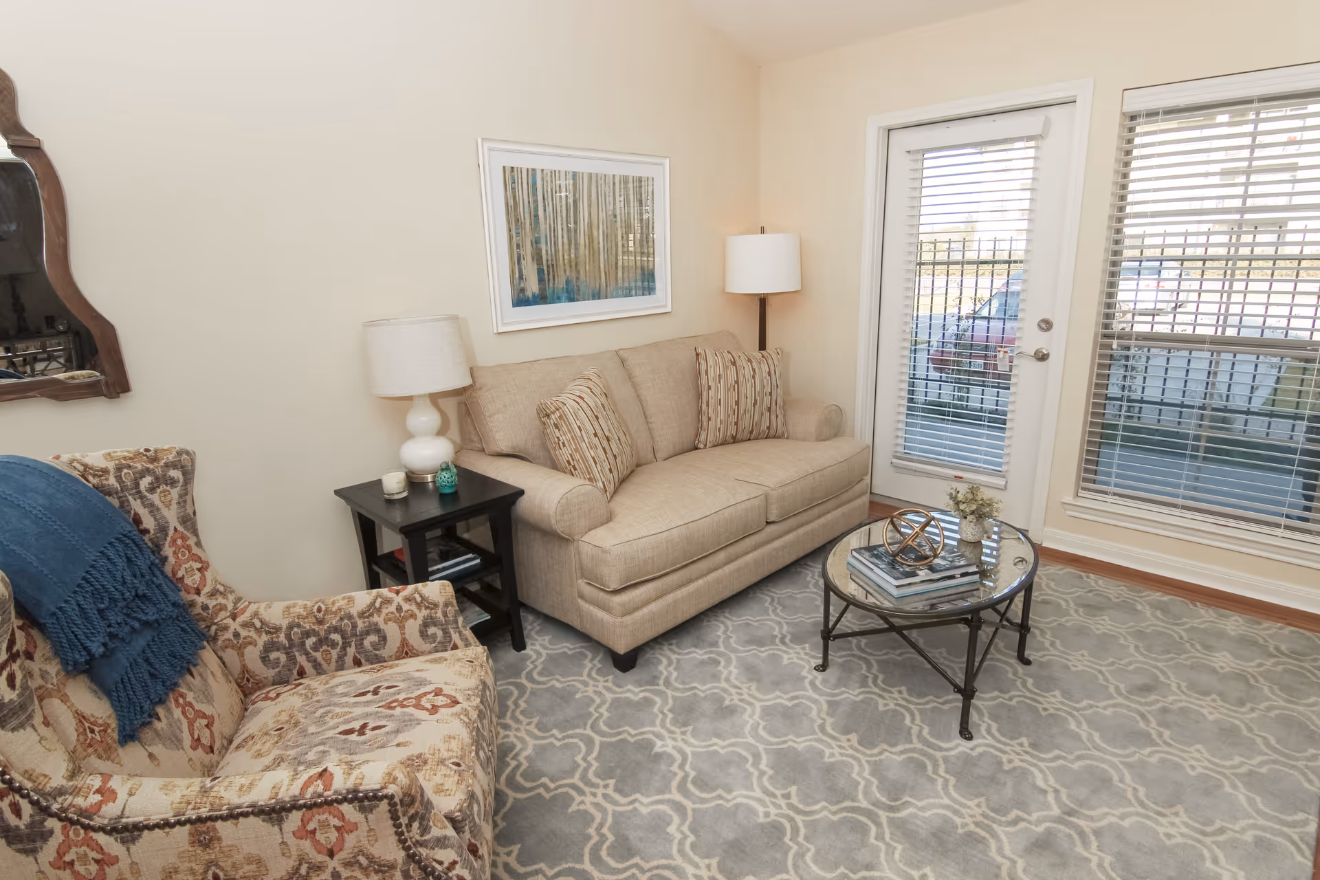A cozy living room with a beige sofa adorned with two patterned pillows, a patterned armchair with a blue throw draped over it, a black side table with a white lamp and small decorative items, a round glass coffee table with books and a decorative object, a floor lamp in the corner, a large window and a glass door with blinds, and a framed artwork on the wall.