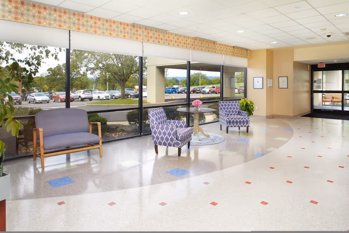 Bright lobby/waiting area with large windows, patterned chairs around a small table, a bench, and a view of the parking lot outside.