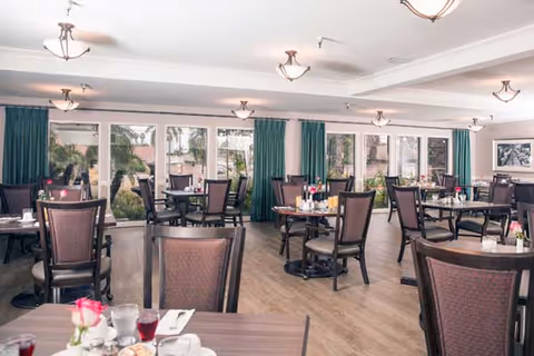 A bright dining room with multiple tables and chairs arranged neatly. Each table is set with glasses, napkins, and small flower vases. Large windows with green curtains allow natural light to fill the room, and there are ceiling lights evenly spaced across the white ceiling.