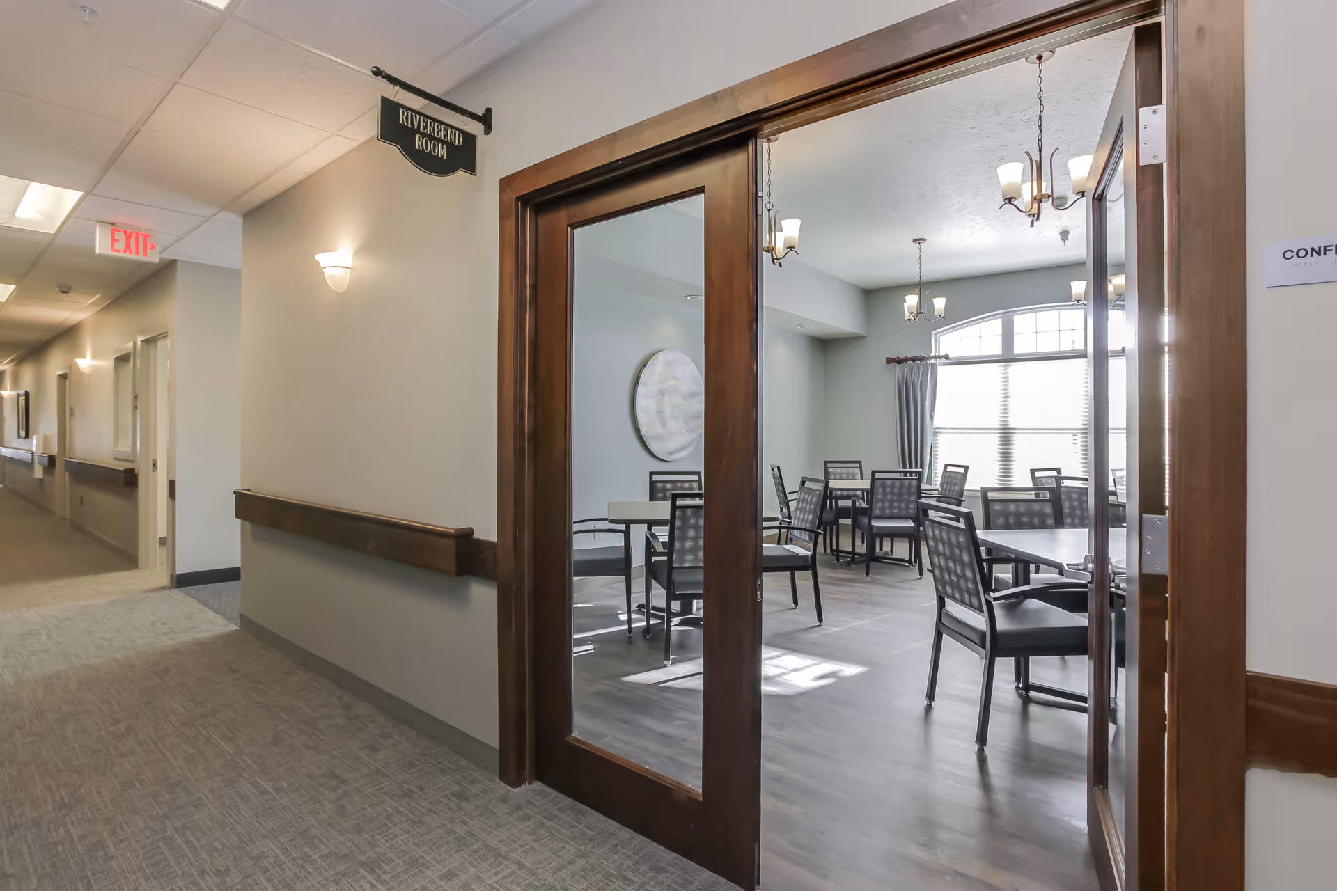 View of a hallway in an assisted living facility leading to a room with glass double doors labeled 'Riverbend Room'. Inside the room are several tables and chairs arranged for group seating, with large windows allowing natural light to fill the space. The hallway has wall-mounted lights and an exit sign visible in the distance.